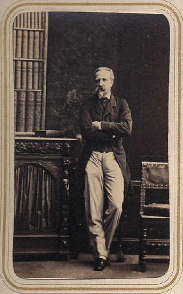Photographie de Henri d'Orléans, duc d'Aumale (1822-1897), en pied, bras croisés, à côté d'une bibliothèque