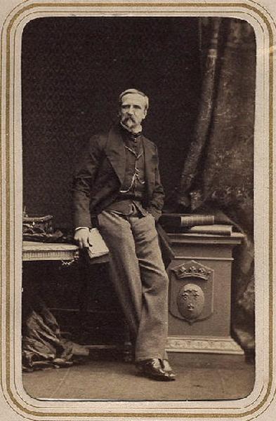 Photographie de Henri d'Orléans, duc d'Aumale (1822-1897), en pied, appuyé sur des livres posés sur un piédestal portant les armes d'Orléans à droite