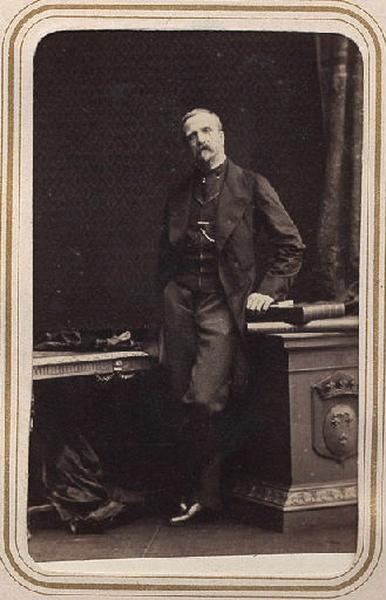 Photographie de Henri d'Orléans, duc d'Aumale (1822-1897), en pied, appuyé sur des livres posés sur un piédestal portant les armes d'Orléans à droite