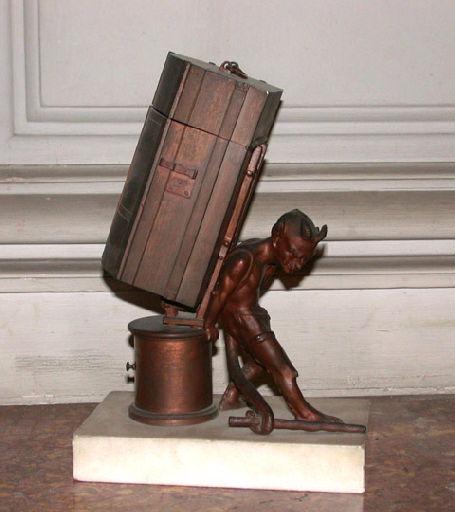 Un porte-allumette représentant un diable portant une malle sur un socle de pierre