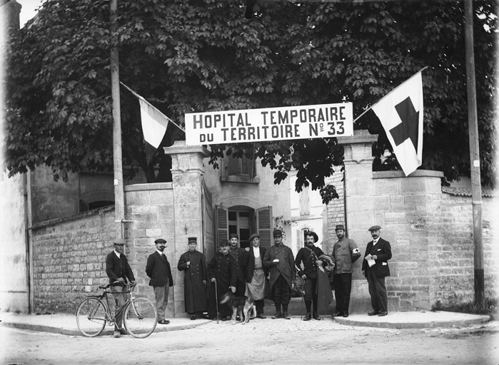 “Hôpital temporaire du territoire n°33” : devant un portail grilles ouvertes, soldats, soignants, drapeau de la Croix-Rouge (titre usuel)