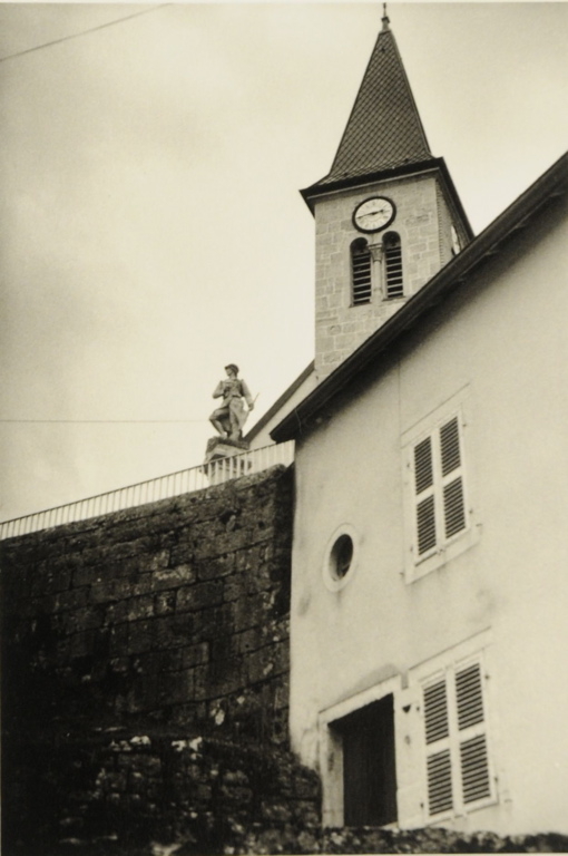 Vue 2009, au premier plan, façade blanche d'une maison et mur de pierre ; soldat d'un monument aux morts et clocher vus di sotto in sù