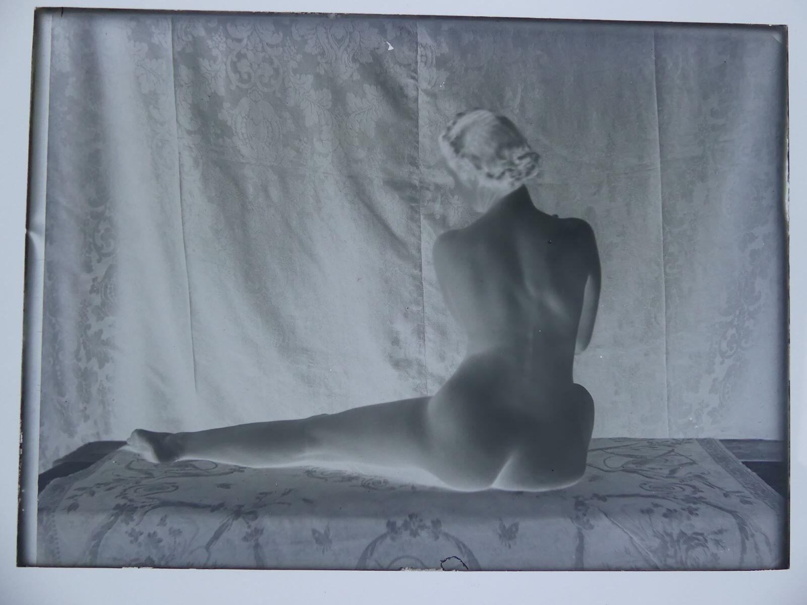 Modèle féminin posant nue, de dos, assise, une jambe repliée, une jambe allongée, les mains croisées sur le buste