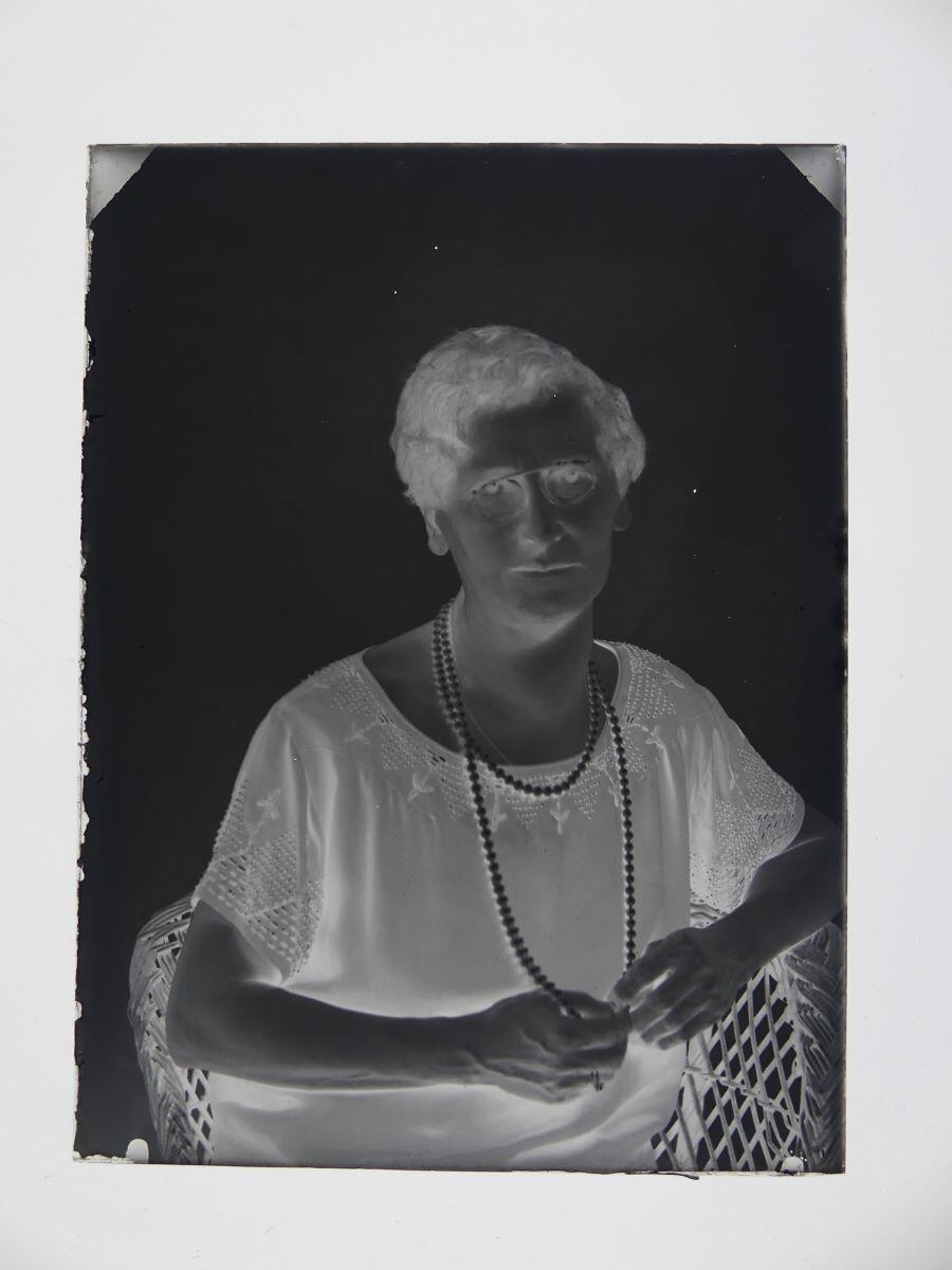 Portrait d'une femme portant des lunettes et un sautoir en perles, assise dans un fauteuil en osier