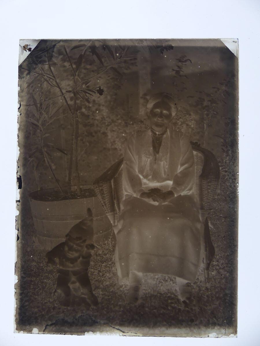 Portrait d'une femme âgée tenant des fleurs et posant dans un fauteuil d'osier, à côté d'un nain de jardin