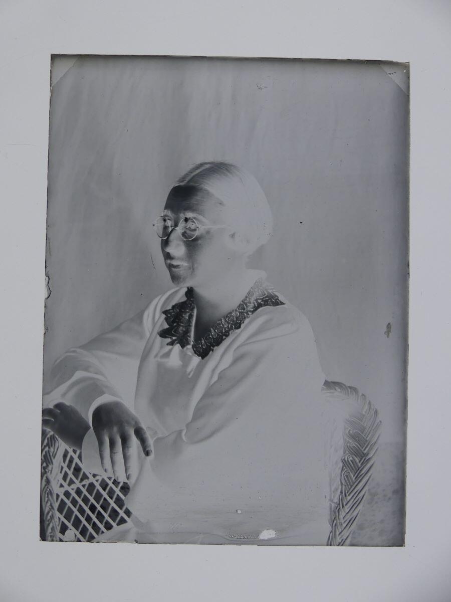 Portrait de femme portant des lunettes, vue de 3/4 droit, assise dans un fauteuil en osier, les mains croisées