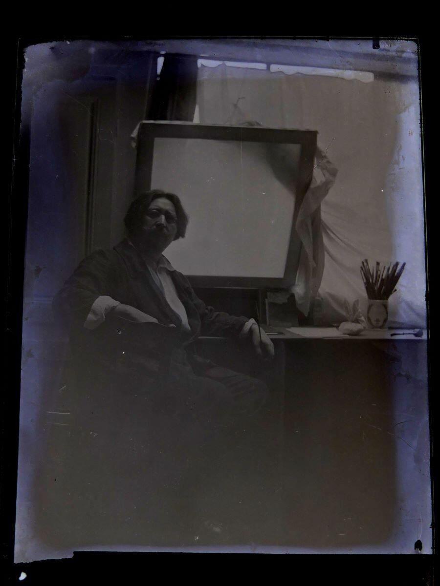 Portrait du peintre Léon Tirode dans son atelier assis devant une table de travail, assis sur la gauche