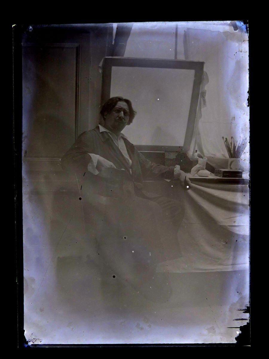 Portrait du peintre Léon Tirode dans son atelier assis devant une table de travail, assis sur la gauche