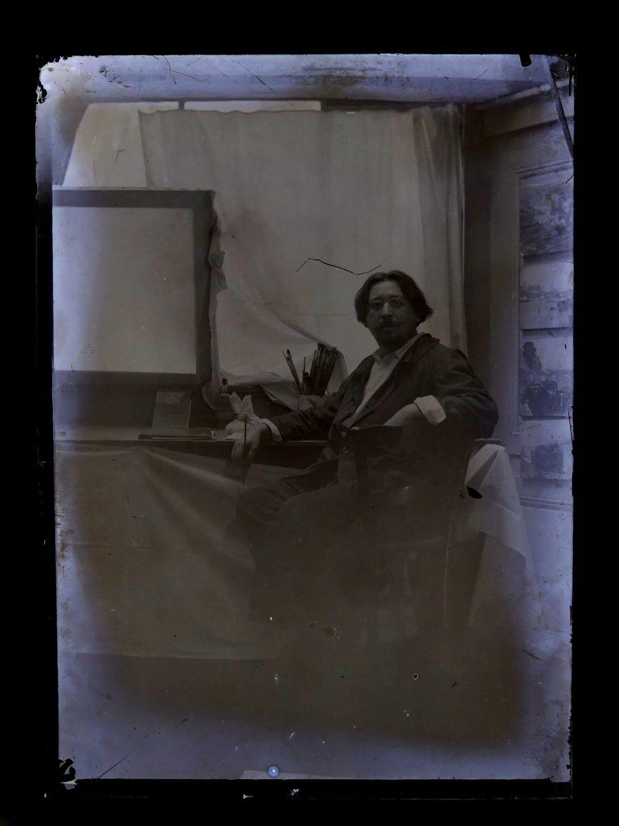 Portrait du peintre Léon Tirode dans son atelier assis devant une table de travail, assis sur la droite