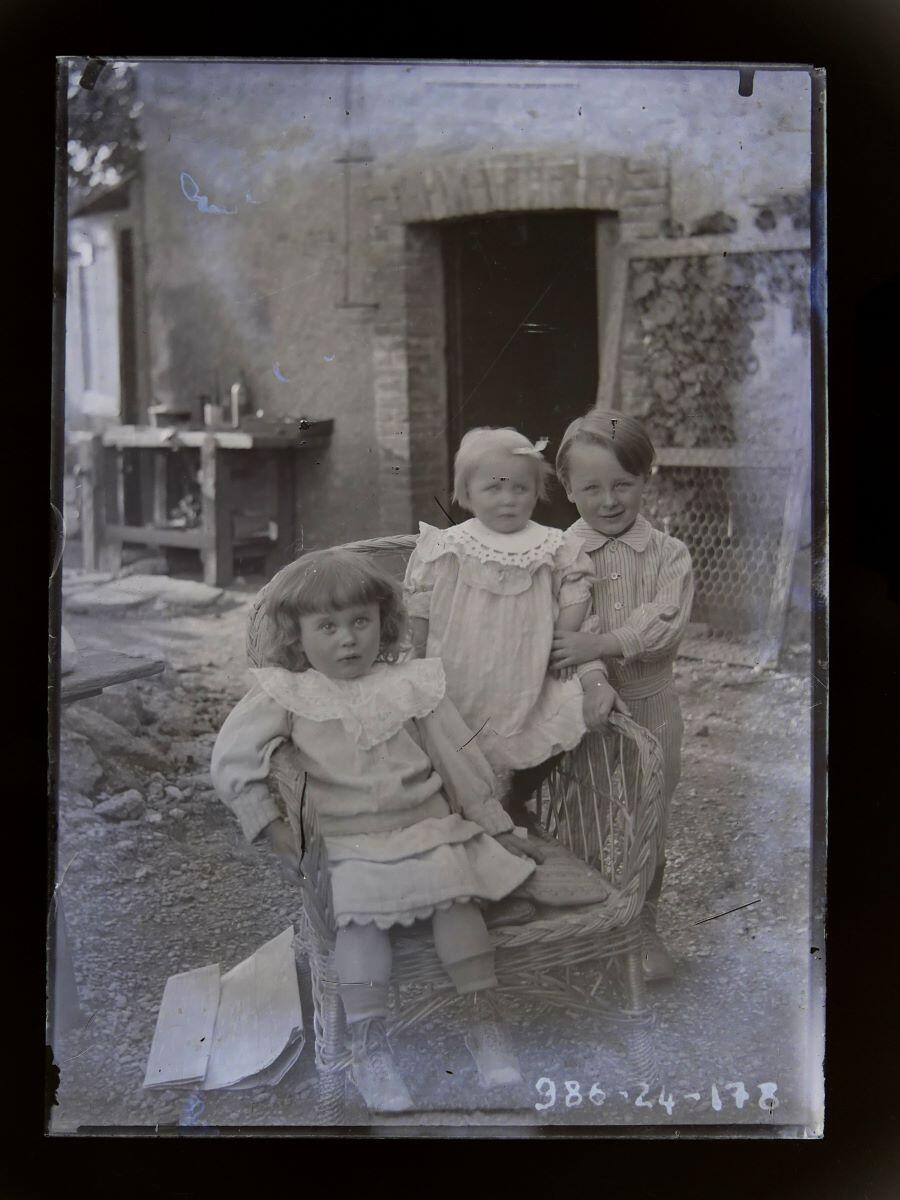 Trois jeunes enfants, dont deux dans un fauteuil en rotin, posant devant une maison